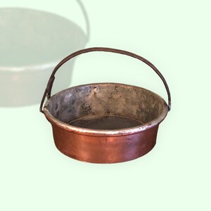 Può includere: Una pentola di rame vintage con un manico in metallo. La pentola ha un aspetto usurato e invecchiato, perfetta per un arredamento rustico.