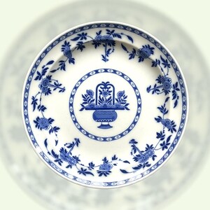 Può includere: Piatto in ceramica bianca con un motivo floreale blu. Il piatto presenta un motivo centrale di un cesto di fiori e un bordo di motivi floreali. Il design è in stile classico e vintage, adatto per servire cibo o come elemento decorativo.