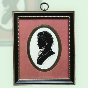 Puede incluir: Una silueta en blanco y negro de un hombre en un marco con un paspart&uacute; ovalado dorado y un fondo rojo. El marco es negro con detalles dorados. El nombre del hombre est&aacute; escrito en cursiva debajo de la silueta.