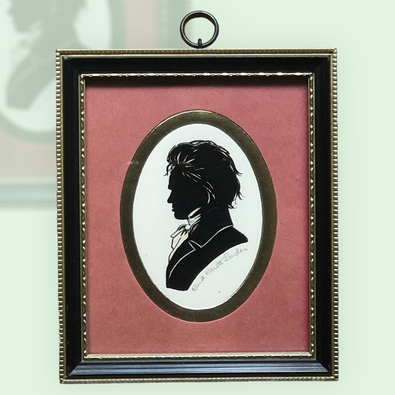 Puede incluir: Una silueta en blanco y negro de un hombre en un marco con un paspart&uacute; ovalado dorado y un fondo rojo. El marco es negro con detalles dorados. El nombre del hombre est&aacute; escrito en cursiva debajo de la silueta.