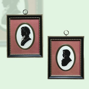 Puede incluir: Dos retratos en silueta enmarcados de personas en negro sobre fondo rojo. Los marcos son negros con adornos dorados. Los retratos son de forma ovalada y est&aacute;n colocados sobre un fondo blanco.