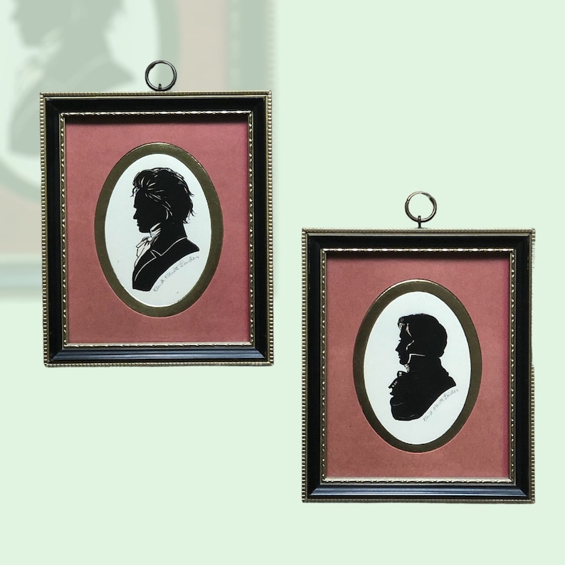 Puede incluir: Dos retratos en silueta enmarcados de personas en negro sobre fondo rojo. Los marcos son negros con adornos dorados. Los retratos son de forma ovalada y est&aacute;n colocados sobre un fondo blanco.
