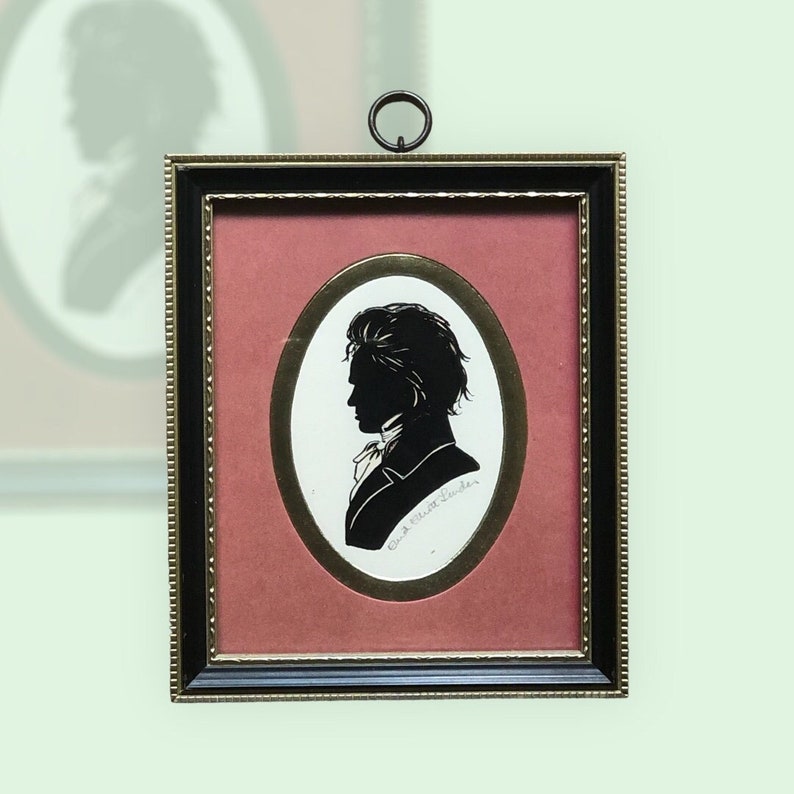 Puede incluir: Una silueta en blanco y negro de un hombre en un &oacute;valo enmarcado. El marco es negro con un borde interior dorado y una montura roja. El nombre del hombre, "Carl Albert Schuler", est&aacute; escrito en cursiva debajo de la silueta.
