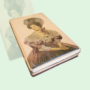 May include: A vintage tin box with a cream background and a picture of a woman in a pink and white dress. The woman is wearing a large hat and has a pearl necklace.