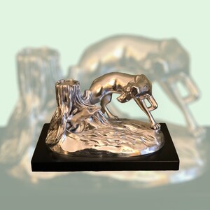 May include: Silver sculpture of a dog chasing a rabbit, set on a black base. The sculpture is detailed with a tree stump and flowing water.