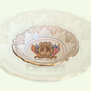 Può includere: Una piccola ciotola in ceramica bianca con bordo dorato e bordo smerlato. La ciotola presenta un'immagine centrale della regina Elisabetta II, bandiere e la scritta "Coronation June 2nd 1953". La bordura è decorata con filigrana dorata.