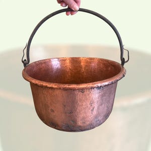 Puede incluir: Un caldero de cobre martillado con un asa de metal oscuro. El caldero tiene una forma redondeada y un borde ligeramente ensanchado. El cobre tiene un color marrón rojizo cálido con algunas áreas más oscuras, lo que sugiere edad y uso.