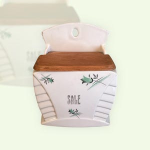 May include: White ceramic wall-mounted salt box with a wooden lid. The box features a curved top and a decorative floral design in teal and grey. The word "SALE" is printed on the front.