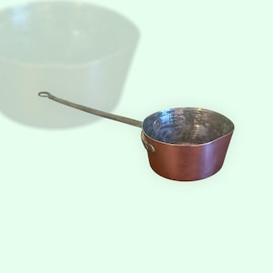 Puede incluir: Una cacerola de cobre vintage con un mango largo y curvo. La cacerola tiene un acabado martillado y está en buen estado.