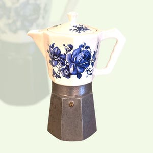 Può includere: Una caffettiera bianca e argentata con un motivo floreale di rose blu. La parte superiore è in ceramica bianca con un motivo floreale blu, e la base è in metallo argentato. Il manico e il coperchio sono bianchi.