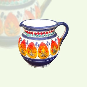 May include: A ceramic pitcher with a blue handle and rim, featuring a white body adorned with vibrant orange, yellow, and red floral designs. The pitcher has a capacity of approximately 1 liter, with a decorative band of red, green, and blue accents.