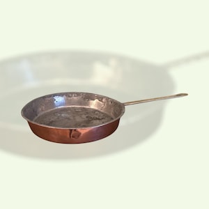 May include: A copper frying pan with a long, silver-colored handle. The pan has a rounded shape with a slightly worn interior. The exterior is a rich, reddish-brown color, typical of copper cookware. The handle is attached with a rivet.