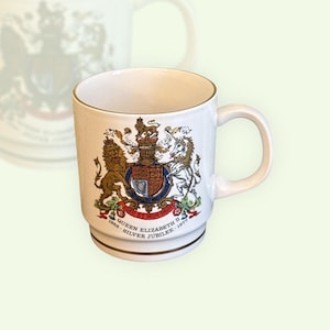 Può includere: Tazza in ceramica bianca con bordo e manico dorati. La tazza presenta un dettagliato stemma con un leone, un unicorno e il testo "Queen Elizabeth II Silver Jubilee 1952-1977".