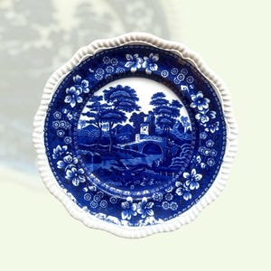 Op de afbeelding: Een blauw en wit decoratief bord met een landschapsscène met een brug, bomen en een gebouw. Het bord heeft een geschulpte rand en bloemmotieven. De rand is wit en het centrale ontwerp is blauw.