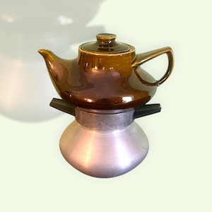 Può includere: Una teiera in ceramica marrone con coperchio abbinato è appoggiata sopra una caffettiera in metallo color argento. La teiera ha un beccuccio e un manico curvi. La caffettiera ha una base arrotondata e una parte superiore piatta.