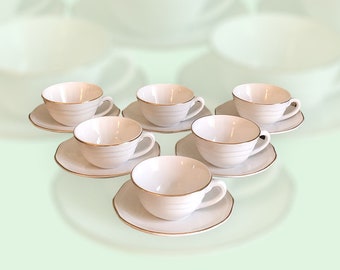 May include: Set of six white teacups and saucers with a gold trim. The cups and saucers are arranged in a semi-circle on a light green background.