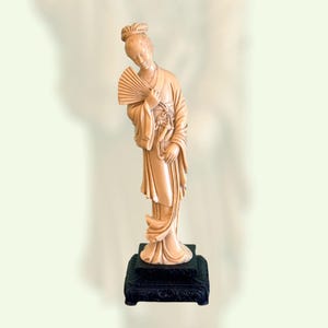 May include: A beige figurine of a woman holding a fan, standing on a black decorative base. The woman is in traditional dress, with her head bowed. The base has an ornate design. The figurine is likely made of resin.