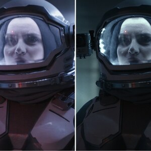 May include: A close-up of a person wearing a space suit helmet with a clear visor. The person's face is visible through the visor. The helmet is dark grey and has a black strap.