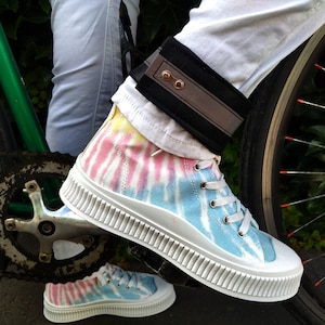 Könnte beinhalten: Ein Paar tie-dye High-Top-Sneaker mit weißen Sohlen und Schnürsenkeln. Die Sneaker werden von einer Person getragen, die Fahrrad fährt. Die Person trägt eine weiße Hose und ein schwarzes reflektierendes Knöchelband.