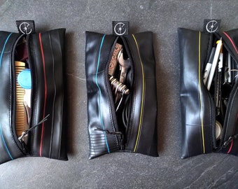 May include: Three black zippered pouches made from recycled bicycle inner tubes. Each pouch has a different colored stripe: red, blue, and yellow. The pouches are filled with various items, such as tools, pens, and pencils.
