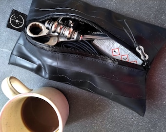 May include: Black zippered pouch made from recycled tire material, partially open to reveal bicycle repair tools. A small tag with a white bicycle wheel logo is attached. A mug of coffee sits nearby.