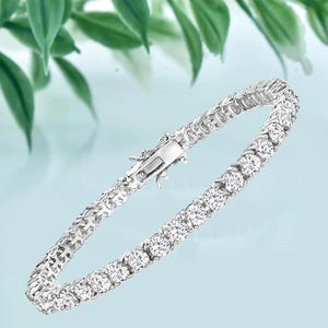 May include: A silver-toned tennis bracelet featuring a continuous row of round, clear, faceted stones set in a metallic frame. The bracelet includes a secure box clasp mechanism with a hinged closure, showcasing a polished finish throughout.