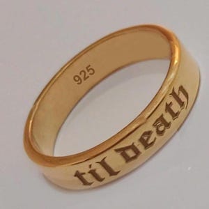 May include: A gold-colored ring with the inscription "til death" in gothic lettering. The ring is marked with "925" on the inside. The ring has a smooth, polished surface.