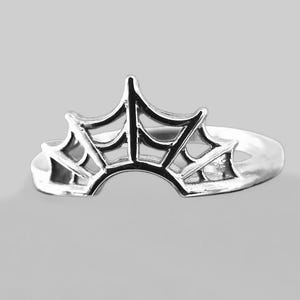 以下が含まれることがあります： クモの巣のデザインが施されたシルバーリング。リングは湾曲したバンドと、上部に詳細なウェブパターンがあります。リングは光沢のある反射性の金属で作られています。