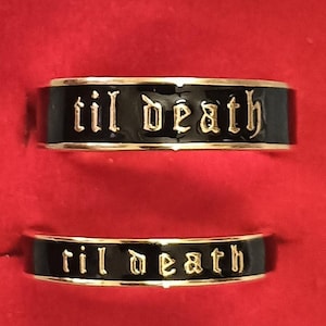 Könnte beinhalten: Zwei schwarze Ringe mit dem Schriftzug "til death" in goldfarbenen Buchstaben. Die Ringe haben einen goldenen Rand und sind vor rotem Hintergrund platziert. Die Ringe sind wahrscheinlich für Paare.