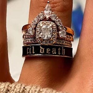 May include: A stack of rings featuring a gold band, a diamond-studded crown-shaped ring, and a black band inscribed with "til death" in white lettering. The rings are worn on a finger, showcasing the intricate details and design.