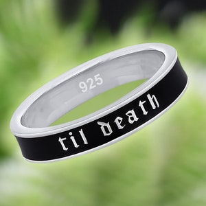 May include: A silver ring with a black band, featuring the inscription "til death" in white gothic lettering. The ring is marked with "925" and has a polished finish, set against a blurred green background.