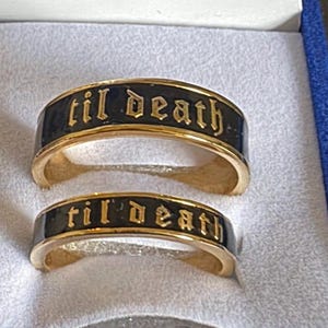 May include: Two gold-colored rings with black bands, each inscribed with the phrase "til death" in an ornate font. The rings are displayed inside a blue velvet ring box, suggesting a theme of commitment or remembrance.