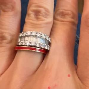 Peut inclure: Une main portant une bague en argent avec une bande rouge et une bande de diamants. La main a deux petites marques rouges sur la paume.