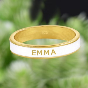 Puede incluir: Un anillo de oro con una banda de esmalte blanco y el nombre "EMMA" grabado en él. El anillo está marcado con "925" en el interior.
