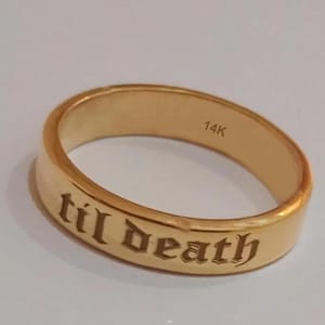 May include: A gold band ring featuring the engraved text 'til death' on the outer surface in a gothic-style font. The interior of the ring is clearly stamped with the text '14K', indicating the gold purity of the metal band.