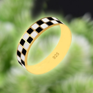 May include: A gold-colored ring with a checkered pattern of black and white squares around the band. The ring is marked with "925" on the inside. The ring is set against a blurred green background.