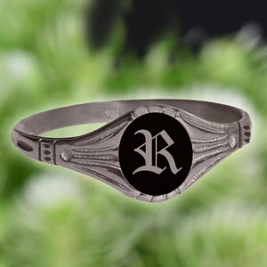 Op de afbeelding: Een zilveren ring met een zwart cirkelvormig middenstuk met een gestileerde letter "R". De ring is gemarkeerd met "925" en heeft decoratieve details op de band. De ring staat tegen een vage groene achtergrond.