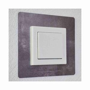Könnte beinhalten: Ein weißer Lichtschalter mit einer grauen, strukturierten Umrandung. Der Schalter ist an einer weißen Wand montiert.