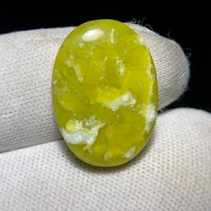 Pode incluir: Uma gema oval cabochão de cor amarela brilhante com veios e redemoinhos brancos. A pedra tem uma superfície lisa e polida.