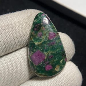 May include: A polished, teardrop-shaped gemstone featuring a deep green matrix with vibrant, irregular patches of ruby. The ruby inclusions are a rich, purplish-red hue, creating a striking contrast against the green background. The stone is held by a gloved hand.