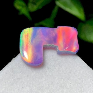 Puede incluir: Una piedra preciosa de ópalo pulida, de forma libre, con un vibrante juego de colores. La piedra muestra tonos de rosa, naranja, morado y verde. El ópalo está colocado sobre una superficie blanca, con un fondo oscuro.