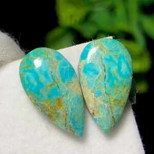 Puede incluir: Dos piedras preciosas turquesas en forma de lágrima con superficie pulida. Las piedras muestran un color azul-verdoso vibrante con patrones de matriz marrón y beige. Las piedras están sobre un fondo blanco.