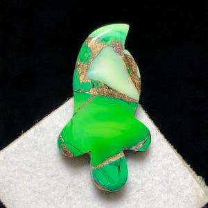 May include: A striking, abstract gemstone in a distinctive shape. The stone has a bright green base with gold-coloured veins and accents. Sections of the stone are a lighter, almost white-green colour. The gemstone is set against a black background.