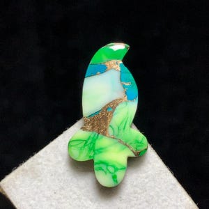 May include: An abstract pendant in the shape of a bird, with green, blue, and white colours. Gold-coloured veins run through the design. The pendant is set against a dark background, which highlights the vibrant colours and intricate details of the piece.