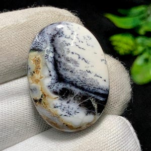 May include: An oval gemstone with a polished finish. The stone displays a white base with detailed patterns in black, grey, and brown. The patterns create a landscape or abstract design. The gemstone is set against a white fabric.