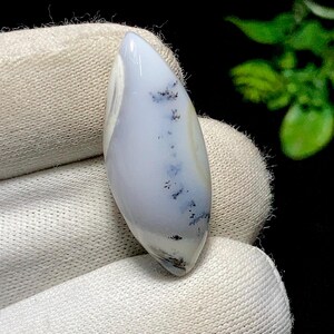 May include: A polished, elongated gemstone with a pale blue and white colour gradient. The stone features delicate black dendrite inclusions, creating a unique pattern. The gemstone is held in a white fabric.