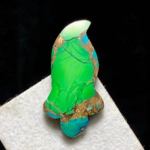 May include: A gemstone in a distinctive shape, displaying vivid green, turquoise, and gold hues. The stone has a polished surface with visible veining and a unique, organic form. The gemstone is set against a white surface with a black background.