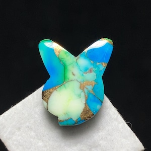 May include: A butterfly-shaped gemstone in shades of blue, green, and white, featuring gold-coloured veins. The stone is displayed on a white surface, set against a black backdrop.