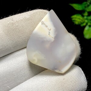 May include: A polished gemstone in a teardrop shape. The stone displays a mix of white, pale yellow, and lavender tones, with delicate dark veining. The gemstone is held in white fabric.