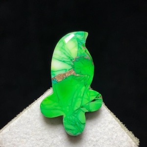May include: A brightly coloured green gemstone with a distinctive shape, displaying a mix of light green, white, and gold-coloured veins. The gemstone is set against a black background, highlighting its bright colours and intricate patterns. Likely used for jewellery making.